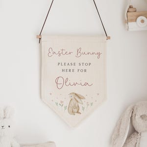 May include: A cream-colored banner with the text "Easter Bunny PLEASE STOP HERE FOR Olivia" and a watercolor illustration of a bunny. The banner is hanging from a wooden rod. A wooden toy camera hangs nearby.