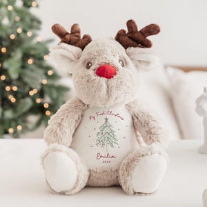 May include: A plush reindeer toy with brown antlers, a red nose, and white accents. The reindeer has a white patch on its chest with the text "My First Christmas", a Christmas tree graphic, and the name "Emilia" with the year "2025".