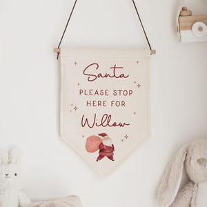 May include: A cream-coloured banner with the text "Santa PLEASE STOP HERE FOR Willow" in burgundy script. A watercolour illustration of Santa Claus carrying a sack is at the bottom. The banner hangs from a wooden rod.