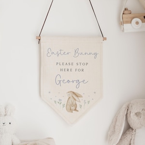 May include: A cream-coloured banner with the text "Easter Bunny, Please Stop Here For George" and a watercolour illustration of a bunny. The banner is suspended from a wooden rod. A wooden toy camera hangs nearby.