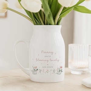 May include: A white ceramic pitcher with a handle, filled with white tulips. The pitcher is decorated with the text "Nanny you're blooming lovely" and floral illustrations. A glass of water and a candle holder are in the background.