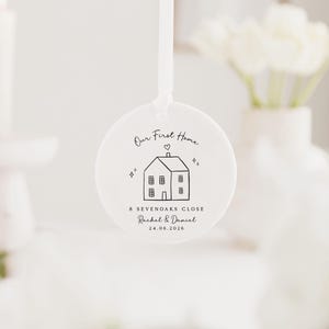 May include: A white, round ornament with a ribbon for hanging. The ornament features a black line drawing of a house with the text "Our First Home", address, and names. The date 24.06.2026 is also printed on the ornament.