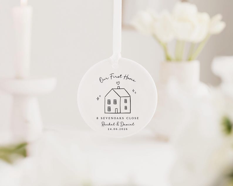 May include: A white, round ornament with a ribbon for hanging. The ornament features a black line drawing of a house with the text "Our First Home", address, and names. The date 24.06.2026 is also printed on the ornament.