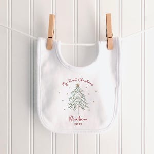 May include: A white baby bib with a Christmas tree design and the text "My First Christmas" in burgundy script. The name "Reuben" and the year "2025" are also printed on the bib. The bib is hanging on a clothesline with wooden clothespins.