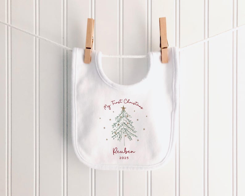May include: A white baby bib with a Christmas tree design and the text "My First Christmas" in burgundy script. The name "Reuben" and the year "2025" are also printed on the bib. The bib is hanging on a clothesline with wooden clothespins.