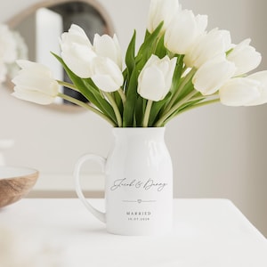 May include: A white ceramic pitcher with a handle, filled with white tulips. The pitcher has the words "Jacob & Danny" and "MARRIED 10.07.2026" printed on it, with a heart symbol. The pitcher is on a white surface.