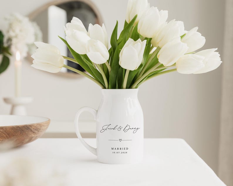 May include: A white ceramic pitcher with a handle, filled with white tulips. The pitcher has the words "Jacob & Danny" and "MARRIED 10.07.2026" printed on it, with a heart symbol. The pitcher is on a white surface.