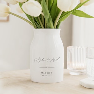 May include: A white ceramic vase with the names "Sophie & Nick" in elegant script, a small heart, and the date "22.06.2026" printed below. The vase holds a bouquet of white tulips. A glass and a candle holder are in the background.