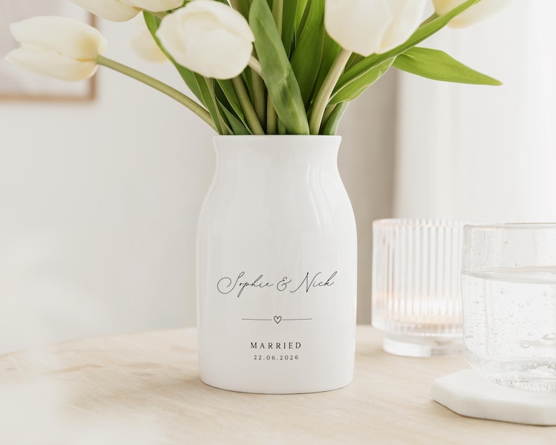 May include: A white ceramic vase with the names "Sophie & Nick" in elegant script, a small heart, and the date "22.06.2026" printed below. The vase holds a bouquet of white tulips. A glass and a candle holder are in the background.