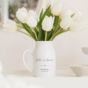 May include: A white ceramic pitcher vase with a handle, filled with white tulips. The vase has the names "Katie & James" and the word "MARRIED" with a date below. A white candle and small bowl are also visible.
