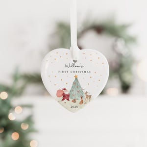 K&ouml;nnte beinhalten: Ein wei&szlig;er, herzf&ouml;rmiger Keramikschmuck, der an einem wei&szlig;en Band h&auml;ngt. Das Ornament zeigt eine gedruckte Illustration von Santa Claus, einem Weihnachtsbaum und einem Reh sowie den Text Willow's FIRST CHRISTMAS und das Jahr 2025.
