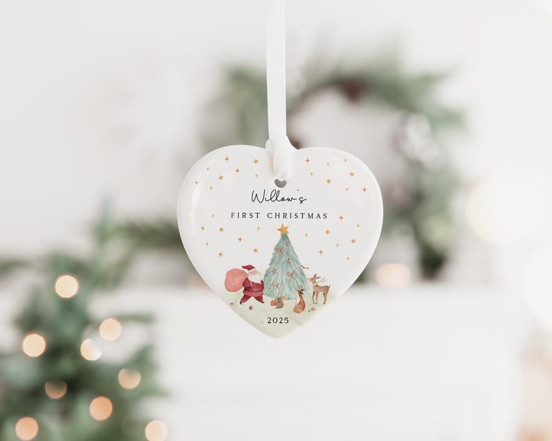 K&ouml;nnte beinhalten: Ein wei&szlig;er, herzf&ouml;rmiger Keramikschmuck, der an einem wei&szlig;en Band h&auml;ngt. Das Ornament zeigt eine gedruckte Illustration von Santa Claus, einem Weihnachtsbaum und einem Reh sowie den Text Willow's FIRST CHRISTMAS und das Jahr 2025.