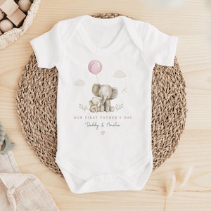 May include: White baby onesie with a watercolor illustration of two elephants, a pink balloon, and the text "OUR FIRST FATHER'S DAY" and "Daddy & Amelia". The onesie is on a woven mat.