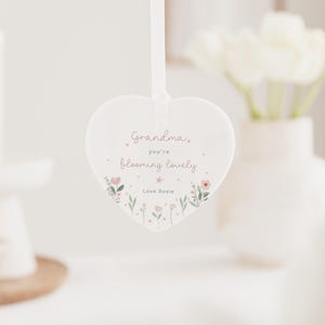 May include: A heart-shaped, white ceramic ornament with a ribbon for hanging. The ornament features the text "Grandma, you're blooming lovely" in pink script, with floral illustrations. The text "Love Rosie" is at the bottom.