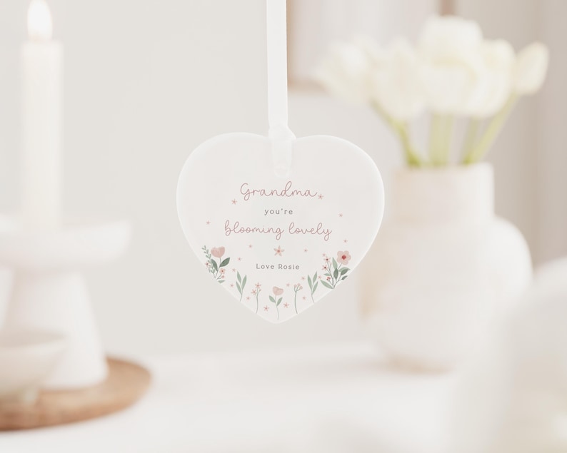 May include: A heart-shaped, white ceramic ornament with a ribbon for hanging. The ornament features the text "Grandma, you're blooming lovely" in pink script, with floral illustrations. The text "Love Rosie" is at the bottom.