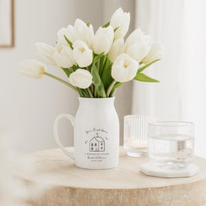 May include: A white ceramic pitcher with a handle, filled with white tulips, sits on a wooden table. The pitcher has a black graphic of a house and the text "Your First Home". A glass of water and a candle holder are also on the table.