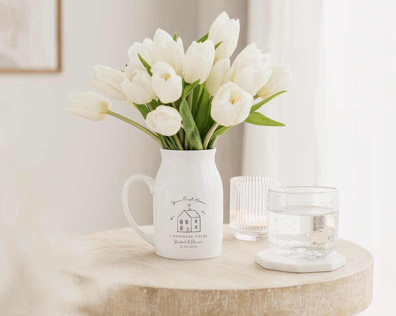 May include: A white ceramic pitcher with a handle, filled with white tulips, sits on a wooden table. The pitcher has a black graphic of a house and the text "Your First Home". A glass of water and a candle holder are also on the table.