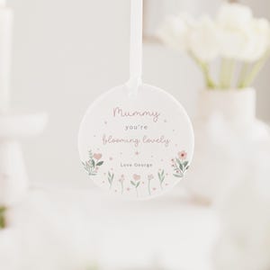 May include: A round, white hanging ornament with the text "Mummy you're blooming lovely" in pink script. The ornament is decorated with small pink flowers and green leaves. A white ribbon is attached for hanging.