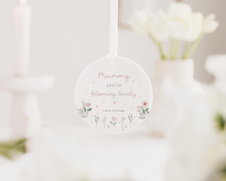 May include: A round, white hanging ornament with the text "Mummy you're blooming lovely" in pink script. The ornament is decorated with small pink flowers and green leaves. A white ribbon is attached for hanging.