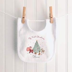 K&ouml;nnte beinhalten: Ein wei&szlig;es L&auml;tzchen mit einem aufgedruckten Weihnachtsbaum, dem Weihnachtsmann, einem Rentier und einem Eichh&ouml;rnchen. Der Text lautet My First Christmas und der Name Oliver. Das L&auml;tzchen h&auml;ngt an zwei h&ouml;lzernen W&auml;scheklammern.
