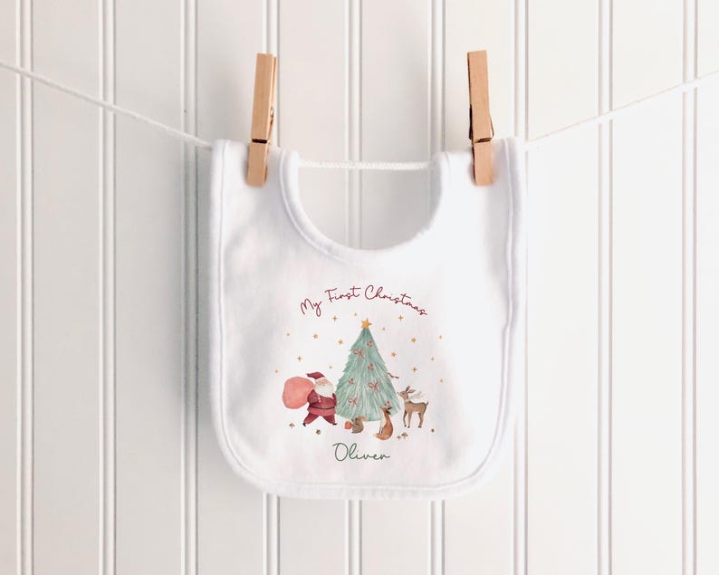 K&ouml;nnte beinhalten: Ein wei&szlig;es L&auml;tzchen mit einem aufgedruckten Weihnachtsbaum, dem Weihnachtsmann, einem Rentier und einem Eichh&ouml;rnchen. Der Text lautet My First Christmas und der Name Oliver. Das L&auml;tzchen h&auml;ngt an zwei h&ouml;lzernen W&auml;scheklammern.