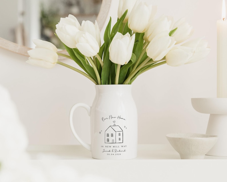 May include: A white ceramic pitcher with a handle, holding a bouquet of white tulips. The pitcher has a black line drawing of a house and the text "Our New Home" and "16 New Mill Way Jacob & Richard 10.04.2024".