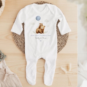 May include: White baby onesie with long sleeves and attached feet. The onesie features a watercolor illustration of two brown bears, one holding a blue balloon. Text on the onesie reads "OUR FIRST FATHER'S DAY" and "Daddy & Oliver".