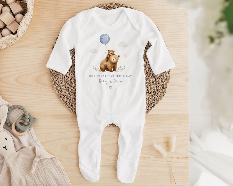 May include: White baby onesie with long sleeves and attached feet. The onesie features a watercolor illustration of two brown bears, one holding a blue balloon. Text on the onesie reads "OUR FIRST FATHER'S DAY" and "Daddy & Oliver".
