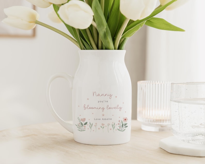 May include: A white ceramic pitcher with a handle, filled with white tulips. The pitcher is decorated with the text "Nanny you're blooming lovely" and floral illustrations. A glass of water and a candle holder are in the background.
