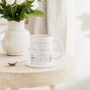 May include: White ceramic mug with the text "Mummy you're blooming lovely" and floral illustrations. The mug sits on a white coaster on a light-colored wooden table. A vase of white flowers is in the background.