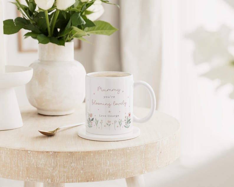May include: White ceramic mug with the text "Mummy you're blooming lovely" and floral illustrations. The mug sits on a white coaster on a light-colored wooden table. A vase of white flowers is in the background.