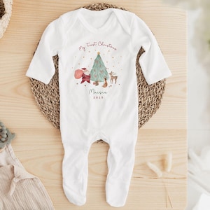 K&ouml;nnte beinhalten: Ein wei&szlig;er lang&auml;rmeliger Baby-Body mit einem festlichen Motiv aus Weihnachtsmann, Weihnachtsbaum und Rentier. Der Text lautet My First Christmas, der Name Maisie und das Jahr 2025 in einer dunkelroten und grauen Schreibschrift.