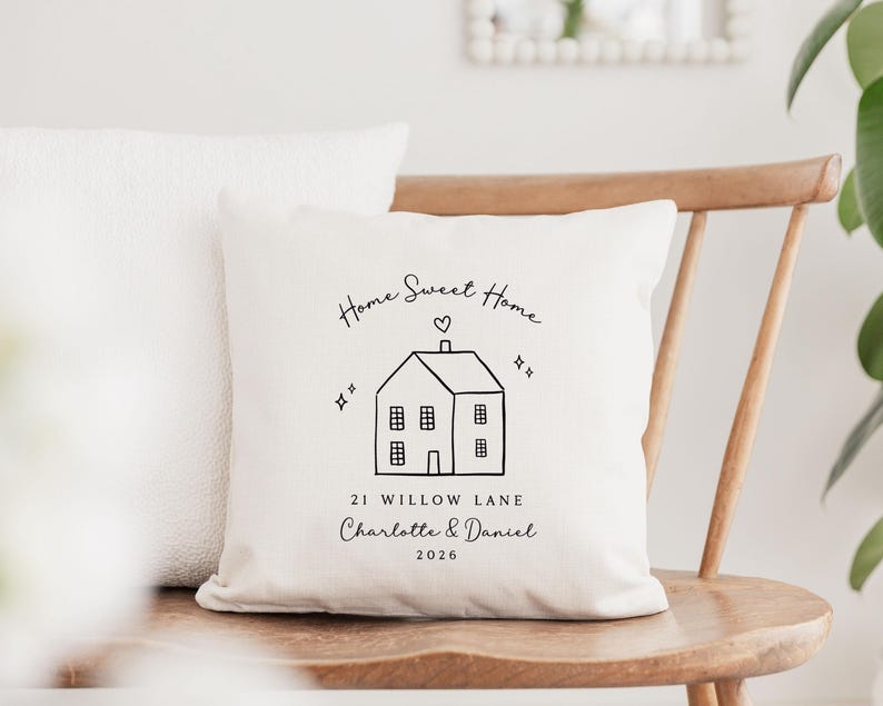 May include: Cream-colored throw pillow with a black line drawing of a house, heart, and the words "Home Sweet Home." Below the house is the text "21 Willow Lane Charlotte & Daniel 2026." Another white pillow is behind it.