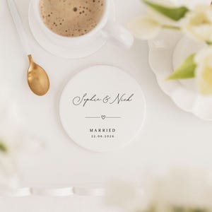 May include: A white ceramic coaster with the names "Sophie & Nick" in elegant script, a heart, and the word "MARRIED" with the date "22.06.2026". A cup of coffee and a gold spoon are nearby, with white flowers in the background.