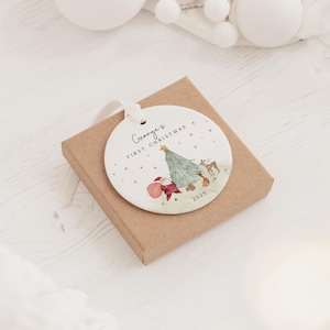 K&ouml;nnte beinhalten: Ein rundes, wei&szlig;es Keramikornament mit wei&szlig;em Band, verziert mit einer Aquarellillustration eines Weihnachtsbaums, eines Rehs und des Weihnachtsmanns. Der Text "George's FIRST CHRISTMAS" und "2025" ist in Schwarz mit goldenen Sternen.