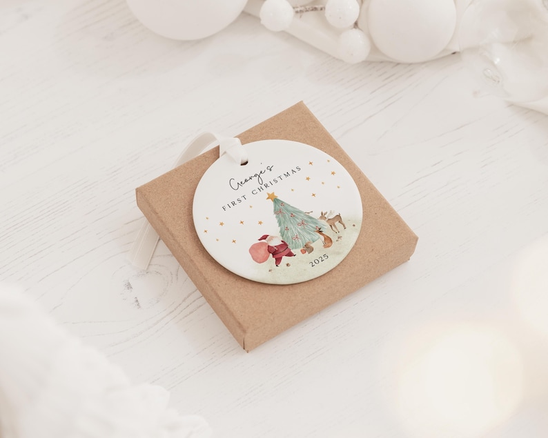 K&ouml;nnte beinhalten: Ein rundes, wei&szlig;es Keramikornament mit wei&szlig;em Band, verziert mit einer Aquarellillustration eines Weihnachtsbaums, eines Rehs und des Weihnachtsmanns. Der Text "George's FIRST CHRISTMAS" und "2025" ist in Schwarz mit goldenen Sternen.