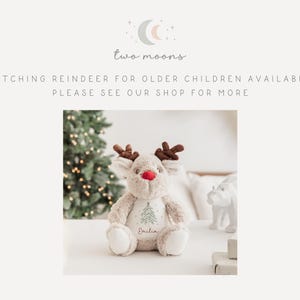 May include: A plush reindeer toy with brown antlers, a red nose, and embroidered details. The reindeer has a green Christmas tree design and the name "Emilia" embroidered on its chest. The text "two moons" is at the top.
