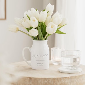 May include: A white ceramic pitcher with a handle, filled with white tulips. The pitcher has the text "Sophie & Nick" and "Married 22.06.2024". A glass of water and a candle holder are on a wooden table.