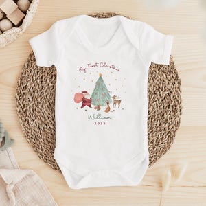 K&ouml;nnte beinhalten: Ein wei&szlig;er kurz&auml;rmeliger Babybody mit einem aufgedruckten Motiv aus Weihnachtsbaum, Weihnachtsmann und einem Reh. Der Text lautet My First Christmas, William und 2025. Das Design enth&auml;lt kleine Sterne und rote Akzente am Baum.