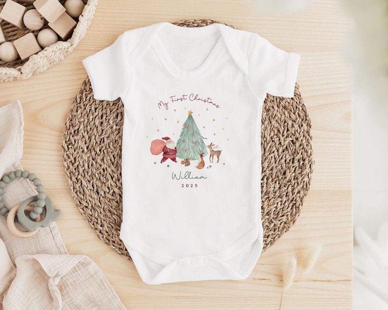 K&ouml;nnte beinhalten: Ein wei&szlig;er kurz&auml;rmeliger Babybody mit einem aufgedruckten Motiv aus Weihnachtsbaum, Weihnachtsmann und einem Reh. Der Text lautet My First Christmas, William und 2025. Das Design enth&auml;lt kleine Sterne und rote Akzente am Baum.