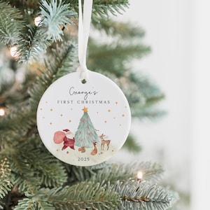 K&ouml;nnte beinhalten: Ein runder, wei&szlig;er Keramik-Weihnachtsbaumschmuck, der an einem wei&szlig;en Band h&auml;ngt. Die Vorderseite zeigt den Text George's FIRST CHRISTMAS und 2025 sowie eine Aquarellillustration vom Weihnachtsmann, einem geschm&uuml;ckten Baum und Waldtieren.