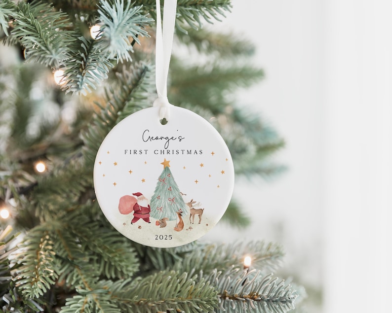 K&ouml;nnte beinhalten: Ein runder, wei&szlig;er Keramik-Weihnachtsbaumschmuck, der an einem wei&szlig;en Band h&auml;ngt. Die Vorderseite zeigt den Text George's FIRST CHRISTMAS und 2025 sowie eine Aquarellillustration vom Weihnachtsmann, einem geschm&uuml;ckten Baum und Waldtieren.