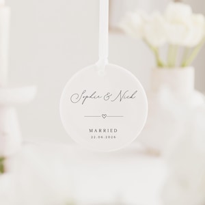 May include: A white, round wedding favor with the names "Sophie & Nick" in elegant script. Below the names is a small heart and a line, with the word "MARRIED" and the date "22.06.2026" printed below. A white ribbon is attached.