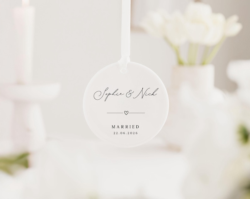 May include: A white, round wedding favor with the names "Sophie & Nick" in elegant script. Below the names is a small heart and a line, with the word "MARRIED" and the date "22.06.2026" printed below. A white ribbon is attached.