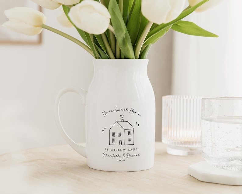 May include: A white ceramic pitcher with a handle, holding white tulips. The pitcher has a black illustration of a house with the words "Home Sweet Home" and a personalized address and names. A glass of water and a candle holder are in the background.