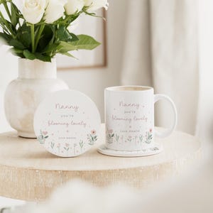 May include: A white ceramic mug and coaster set with the text "Nanny you're blooming lovely" in a delicate script, accompanied by floral illustrations. A vase of white roses sits nearby, creating a soft, inviting scene.