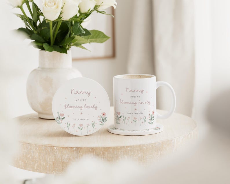 May include: A white ceramic mug and coaster set with the text "Nanny you're blooming lovely" in a delicate script, accompanied by floral illustrations. A vase of white roses sits nearby, creating a soft, inviting scene.