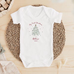 May include: White baby onesie with a Christmas tree design and the text "My First Christmas" and "Arthur 2025". The onesie is on a woven mat with wooden toys and a teething ring nearby. A festive holiday baby outfit.