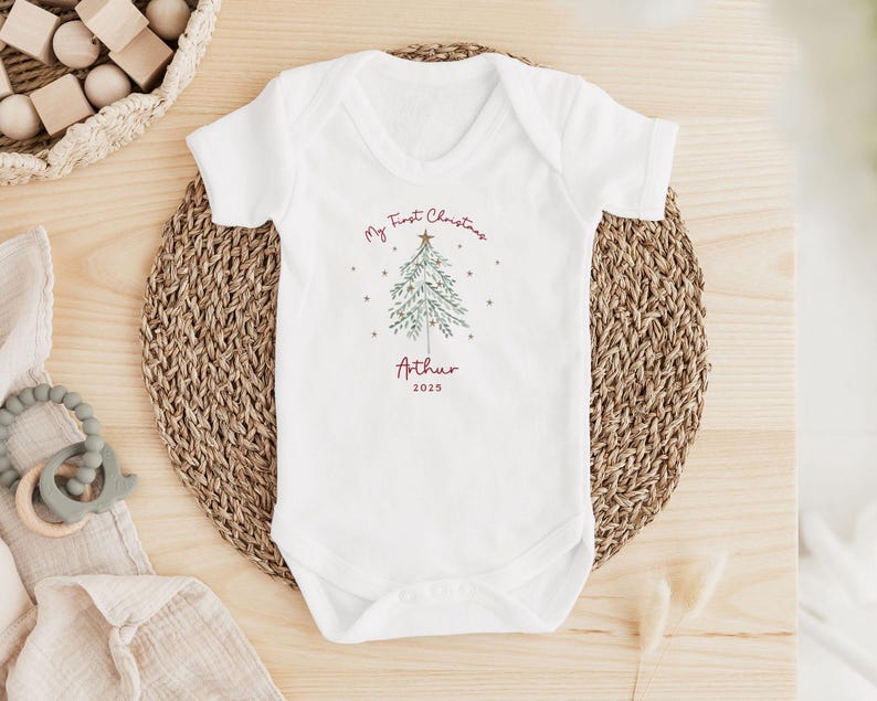 May include: White baby onesie with a Christmas tree design and the text "My First Christmas" and "Arthur 2025". The onesie is on a woven mat with wooden toys and a teething ring nearby. A festive holiday baby outfit.