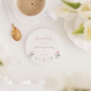 May include: A white ceramic coaster with the text "Grandma you're blooming lovely" in pink script, surrounded by floral illustrations. A gold spoon and a cup of coffee are visible in the background.